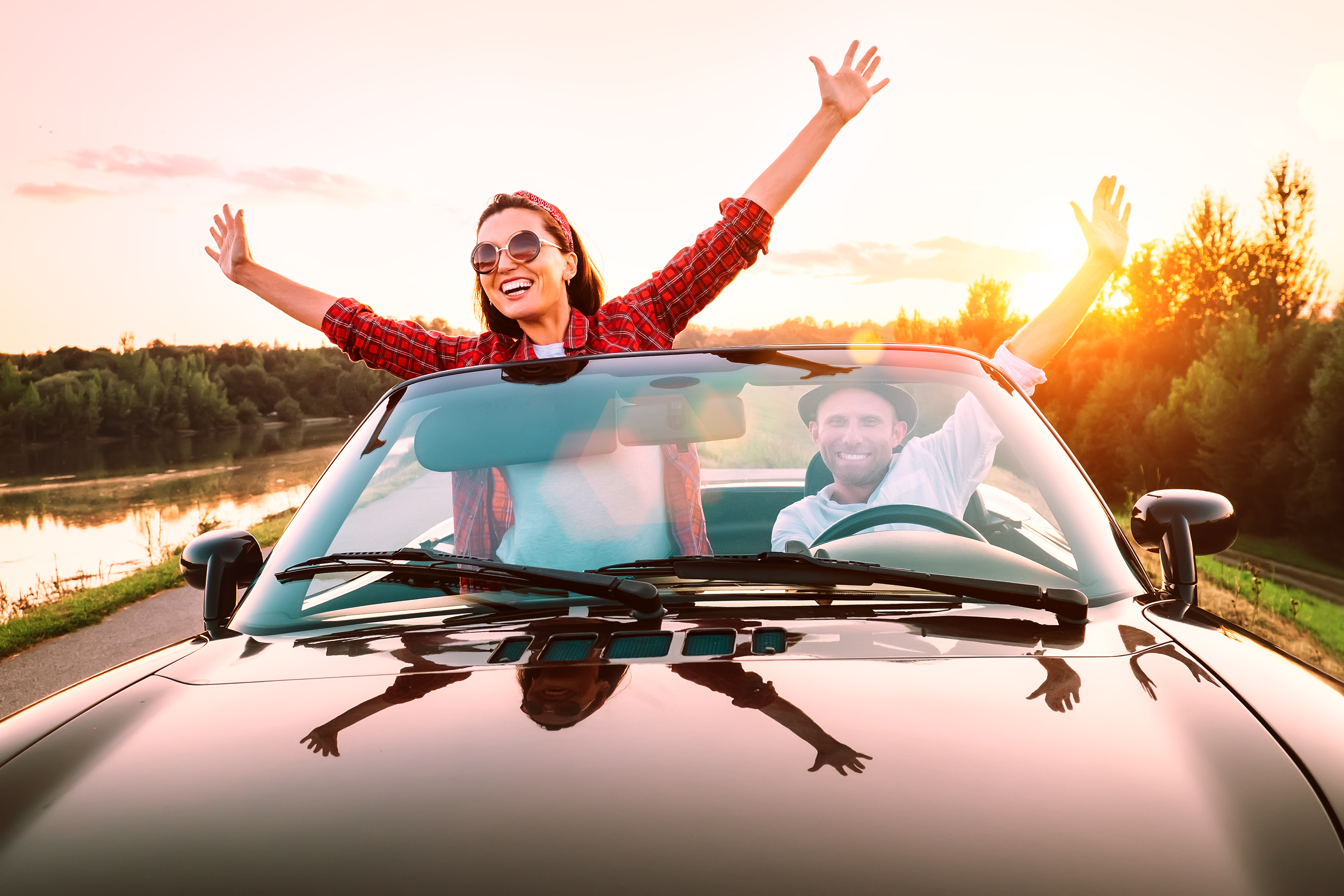 Cabriolet mit zwei Personen. Beide heben die Arme in die Luft. Sonnenuntergang im Hintergrund.